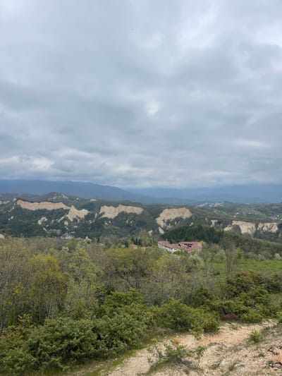 This is an outdoor, daytime shot of a mountainous landscape under a cloudy sky, likely taken from a high vantage point. The view consists of a deep valley with textured rocky terrain interspersed with green vegetation and trees. In the middle ground, there appears to be a building with a reddish-brown roof surrounded by more greenery. The terrain extends into the distance towards the right side of the frame. On the far left, the sky is overcast with a thick layer of clouds. The composition gives a sense of depth and scale to the landscape. The location appears to be Rozhen, Bulgaria, based on the terrain and vegetation.