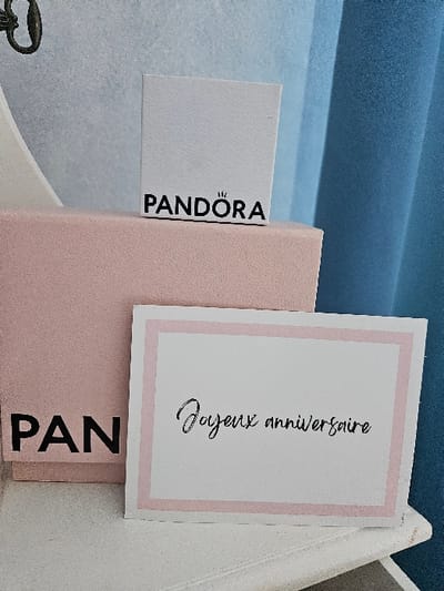 The image displays a close-up, static indoor scene in Villeneuve-Saint-Georges, France. The exact specific location within the city cannot be determined from the visual information provided. The primary subjects are three gift-related items arranged on a white, slightly textured surface, possibly a shelf or table.
From left to right and front to back, the objects are:
1. A white rectangular greeting card with a light pink border. Black script text on the card reads "Joyeux anniversaire" (Happy Birthday). The card is angled, leaning against a larger box.
2. A light pink, rectangular Pandora box with a textured surface, partially visible behind the greeting card. The letters "PAN" from the "PANDORA" logo are visible in black on its front side.
3. A smaller, white rectangular Pandora box standing upright on top of the larger pink box. The complete "PANDORA" logo, with a crown symbol above the 'O', is visible in black on its front.
The background consists of several elements. To the upper left, a dark, curvilinear object, possibly a metallic hook or ornamental part of a mirror/furniture, is partially visible against a light grey or pale blue textured surface. To the right, a set of vertically folded blue fabric, likely curtains, provides a backdrop, with variations in shade from lighter to darker blue. No people are present, and no actions or interactions are depicted. The lighting is uniform, suggesting an indoor setting.