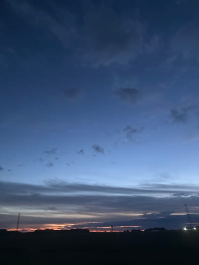 The image shows a dark silhouette of trees and a field against a twilight sky over the Грозаўскі сельскі Савет, Belarus. The sky transitions from a deep blue at the top to lighter shades of blue and orange near the horizon, with scattered clouds. There are faint lights visible on the right side of the horizon, possibly from buildings or streetlights. A thin crescent moon is visible in the sky above the horizon on the left.