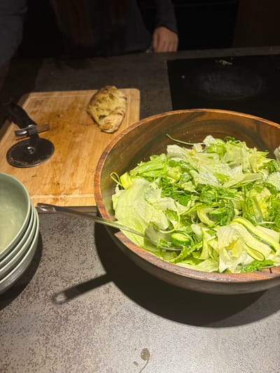 An overhead view captures a kitchen counter scene in Ness Ziona, Israel, under ambient lighting. In the foreground, a dark grey, speckled countertop displays a stack of four light green ceramic bowls on the far left. To their right, a large, dark brown wooden bowl is filled with a green salad, comprising light green lettuce, darker green arugula, and thinly sliced cucumber, with a metal serving spoon partially submerged. Adjacent to the salad bowl, on a light brown wooden cutting board, rests a single, golden-brown, savory pastry or calzone with green herb speckles. A pizza cutter with a black handle is positioned near the pastry on the cutting board. In the upper background, the torso and part of the left arm of a person wearing a dark long-sleeved shirt are visible, positioned behind the counter and to the left. To the right of the cutting board, a dark, reflective cooktop with faint circular burner outlines is partially visible.