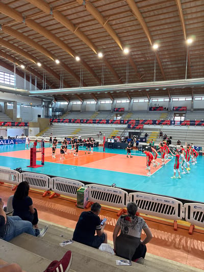 The image depicts a volleyball match taking place in an indoor gymnasium in Cisterna di Latina, Italy. The gymnasium features a high, vaulted wooden ceiling with exposed beams and lighting fixtures. The court is divided by a net, with two teams of young male athletes in distinct uniforms competing. One team is wearing red and white uniforms, and the other is in dark blue and orange uniforms. Spectators are seated in tiered bleachers surrounding the court. Some spectators are visible in the foreground, sitting on the steps. Banners with the text "CEV U18 Volleyball European Championship 2026" and "Cisterna di Latina" are displayed prominently in the background. The court itself is marked with colored lines, indicating the boundaries of play. The scene captures a moment of athletic competition within a sports facility.