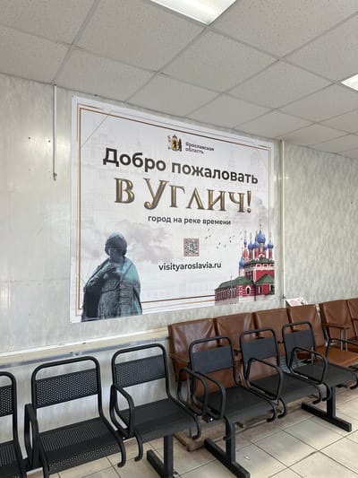 A large banner hangs on a wall, featuring an image of the city of Uglich, Russia, with the text "Добро пожаловать В УГЛИЧ!" (Welcome to Uglich!) and "город на реке времени" (city on the river of time). The banner also displays the website "visityaroslavia.ru" and a QR code. In the foreground, several rows of chairs are visible. The chairs in the front are black, metal chairs with perforated seats and backs, and curved armrests. Behind these are rows of brown leather chairs. The environment appears to be an indoor waiting area.