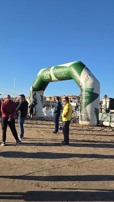 The image captures an outdoor event on the sandy beach area of Complejo Residencial De Vera, Spain, under a clear blue sky. Central to the scene is a large, inflated archway, primarily green and white with geometric designs, displaying the word "CANCER" on its left side and the "aecc" logo with the slogan "Tu Lucha es Nuestra Victoria" on its right.
In the midground, a group of adult males is present. On the left, a man in a dark red hoodie and black pants stands with his hands clasped behind his back, facing right. Adjacent to him is another man in a black hoodie and blue jeans with his arms crossed. To the right, a man in a yellow t-shirt and dark track pants stands with his hands on his hips, facing left, while another man in a dark jacket and blue jeans stands behind him. A person in a dark wetsuit is partially visible beneath the arch, facing away.
The ground consists of light-colored sand, marked by several long, distinct shadows, indicating a low sun angle. In the background, a row of multi-story buildings and palm trees is visible along the horizon, suggesting a developed coastal area. Event equipment, including a black speaker on a tripod and white barriers, is discernible on the right side of the frame.