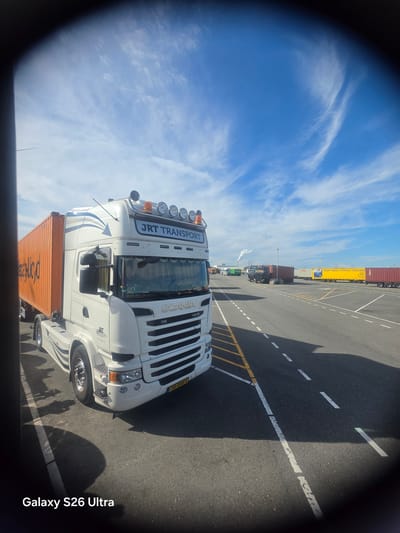 A white Scania truck with "JRT TRANSPORT" branding is parked on a paved area, likely a port or industrial yard, under a bright blue sky with wispy white clouds. The truck is hauling an orange shipping container. In the background, other trucks and trailers are visible, including a yellow trailer with the text "OLOF MEGA TRAVEL TRAILERS". A plume of white smoke rises in the distance, suggesting industrial activity. The lighting indicates it is daytime. The scene appears to be at Maasvlakte in Rotterdam, Netherlands, given the context. The overall impression is one of a busy logistics or transport hub on a clear day. The bottom right corner of the image displays the text "Galaxy S26 Ultra".
