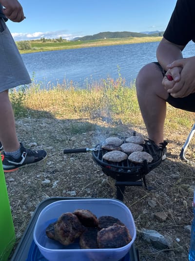 The image depicts an outdoor picnic scene beside a body of water, likely a lake, under a clear blue sky with some scattered clouds. In the foreground, a small black grill is set up on the rocky, dry ground, cooking several hamburger patties. To the left, the legs of a person wearing grey shorts and athletic shoes are visible. To the right, another person's legs, clad in black shorts, are shown, with their hands clasped, holding a small red object, possibly a lighter or a tool. A blue plastic container filled with cooked hamburger patties sits on a grey plastic mat in the lower left corner of the image. The background features dry grass and vegetation, with a serene view of the water and rolling hills beyond. The overall impression is a relaxed, casual gathering for a barbecue in a natural setting. The location is identified as Drenkovo, Bulgaria.