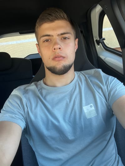 This is a portrait-style media file featuring a young man from the chest up. He is looking directly at the camera with a neutral expression. He has short, dark hair, brown eyes, and a short beard and mustache. He is wearing a light gray t-shirt with a small, white graphic on the left side of his chest depicting a square containing stylized waves and a circular object. The background suggests he is inside a vehicle, possibly a car, with a window visible to his left. The interior of the vehicle appears to be black and gray. The overall lighting and clear view of the man suggest it is daytime. The provided location context of Ustka, Poland, places this image in a coastal town.