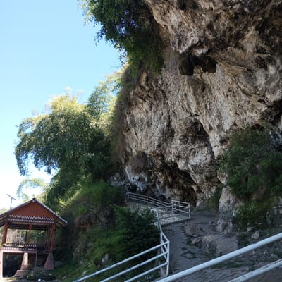 The image depicts an outdoor scene at a natural cave entrance in Kalalengkio, Indonesia. The foreground features a stone pathway with white metal railings, leading towards the cave. The pathway is partially paved with irregular stone slabs and scattered rocks. To the left, a traditional wooden structure with a distinctive patterned roof and white decorative trim stands prominently. This structure appears to be a pavilion or shelter. Lush green vegetation, including trees and bushes, surrounds the pathway and the cave entrance. The cave itself is large and imposing, with a rough, textured rock face that extends upwards. The sky is clear and blue. The scene conveys a sense of natural beauty and potentially a tourist attraction.