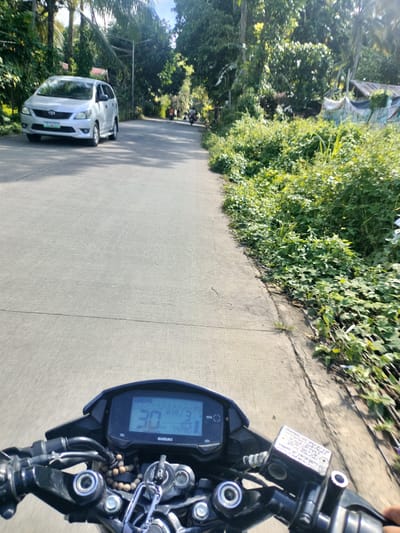 The image shows a first-person perspective from a motorcycle traveling on a paved road. The road is bordered by lush green vegetation on the right side and trees on both sides. A silver car is visible in the distance on the left side of the road. The motorcycle's dashboard displays a speed of 30, indicating the vehicle's current velocity. The scene appears to be in a rural or suburban area, likely in Kidapawan City, Philippines, given the presence of tropical foliage and the style of vehicles. The lighting suggests it is daytime, with sunlight filtering through the trees.