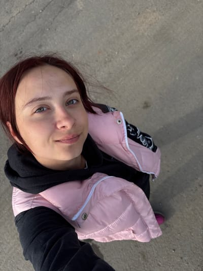 A woman with auburn hair and green eyes is taking a selfie outdoors. She is wearing a black hooded sweatshirt and a pink puffy vest with white trim. She is standing on a gray concrete surface. In the background, a small portion of a pink shoe is visible. There is no indication in the image of the location being Krilovo, Russia.