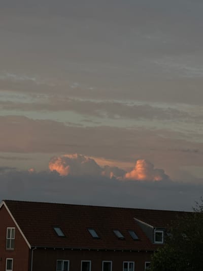 The image shows a rooftop and a dusky sky with illuminated clouds. The rooftop is made of red tiles and features several dormer windows and skylights. A brick building with white-framed windows is visible on the left side. On the right, a tree with green foliage partially obscures the building and sky. The sky is filled with layered clouds, with some in the middle catching an orange-pink light, suggesting it is either sunrise or sunset. The overall ambiance is serene and muted. This scene could be located in any residential area of Roskilde, Denmark, given the architectural style of the building and the common presence of such rooftops in Danish cities. There are no people visible in the image, and no specific actions are taking place.