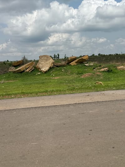 The image depicts an outdoor scene along a paved road in Kuka, Nigeria. The camera is positioned at an angle, with the road occupying the right side of the frame and a grassy embankment on the left. The embankment is dotted with several large, rough-hewn rocks, some of which appear to be flat and stacked. Sparse vegetation, including some trees in the distance, can be seen beyond the rocks. The sky is filled with dramatic, grey-tinged clouds, suggesting a potentially overcast or changing weather condition.

No people are visible in the image. The primary objects are the rocks and the road. The scene appears to be a rural or semi-rural roadside setting. The time of day is not definitively ascertainable, but the lighting suggests it is daytime, likely afternoon given the cloud cover. There is no indication of a specific event taking place; the scene is static and natural.

A notable detail is some faint white markings on the surface of one of the larger rocks, which could be text or symbols, but they are too indistinct to read. The overall impression is one of a natural landscape beside a man-made road, under a cloudy sky.