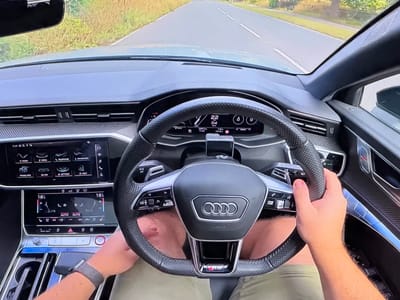 The image shows a first-person perspective from inside a car, specifically an Audi, as it drives down a road. The driver's hands are on the steering wheel, and the dashboard displays are visible, showing navigation and climate control information. The road is paved and flanked by green vegetation. The interior of the car features modern digital displays and carbon fiber accents. There is no visible indication of the car being in Bezden, Bulgaria.