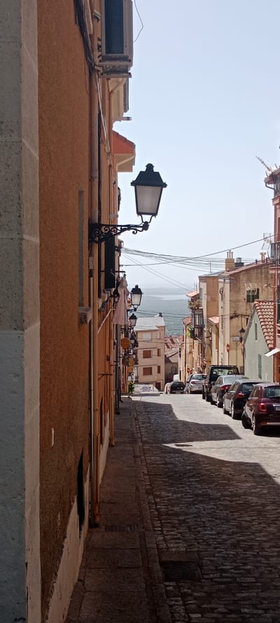 The image depicts a narrow, cobblestone street in San Lorenzo de El Escorial, Spain, ascending a gentle incline. The street is lined with buildings constructed of stone and stucco, with terracotta tiled roofs. Several cars are parked along the right side of the street, casting long shadows due to the bright, clear daytime sky. On the left side of the street, a building with an orange-brown stucco facade features a prominent black wrought-iron street lamp attached to its side. Further down the street, additional street lamps are visible. In the background, a body of water and a distant hazy landscape are discernible. The overall scene conveys a sense of a historic European town on a sunny day.