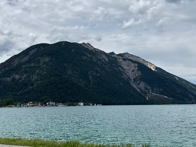 The image shows a wide shot of the Achensee lake in Pertisau, Austria, on an overcast day. In the foreground, the turquoise water of the lake is visible, with ripples on its surface. The shore is lined with green grass and a gravel path. Across the lake, a small town is nestled at the base of a large, dark green, tree-covered mountain. The town consists of several buildings, including hotels and houses, with some boats docked along the waterfront. The mountain dominates the background, with a rocky, lighter-colored section visible on its slope. The sky is filled with gray clouds.