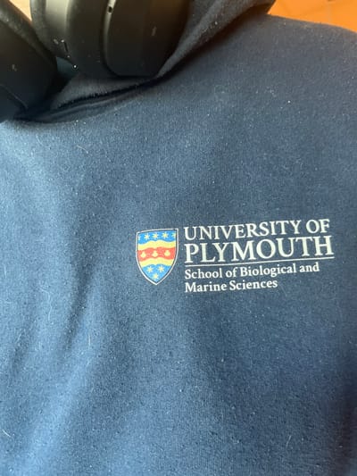 An overhead shot captured at an angle displays a dark blue fabric surface, likely a sweatshirt or hoodie, adorned with the branding of the University of Plymouth. The fabric shows minor pilling and lint, indicating some wear. Text printed in light beige or white is oriented vertically along the right side of the frame, reading "UNIVERSITY OF PLYMOUTH" in a larger, bold font, and beneath it, "School of Biological and Marine Sciences" in a smaller font. To the left of the text, a colorful crest or shield logo is visible, featuring red and yellow wavy stripes, a blue section with white stars, and a solid red section. In the bottom left corner, partially obscured, is a black over-ear headphone, suggesting an indoor setting. The image itself does not provide any specific cues for time of day, weather, or the provided location context of Dorno, Italy. No people are visible.