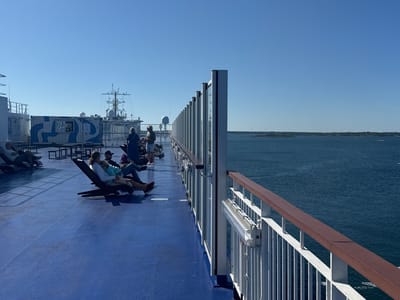 The photograph captures an exterior deck of a ferryboat sailing on a clear, sunny day. The deck is painted a vibrant blue, with white railings and a wooden handrail running along the side. Several people are seated on lounge chairs, facing towards the water, and engaged in various activities like reading or relaxing.
In the background, a large white building structure with blue patterns, possibly a shipping container, is visible on the deck. Further beyond that, the superstructure of the ferry, including antennas and navigational equipment, is apparent. The ferry is navigating through a body of calm, deep blue water, with a distant shoreline dotted with trees and wind turbines, suggesting a coastal landscape. The sky is a clear, cloudless blue.
The image has been identified as being taken in Karlskrona, Sweden, a city known for its naval history and archipelago. The ferry is likely traveling between islands or to the mainland, showcasing a typical scene of maritime transport in the region.