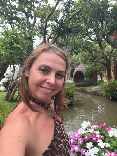 A woman with shoulder-length brown hair smiles at the camera in a selfie. She is wearing a leopard-print bikini top and a black choker necklace. Behind her, a small pond is surrounded by lush greenery and trees. A woven pod-shaped chair hangs from a tree branch over the pond. In the background, a large white bear sculpture with a cup is visible. The scene is outdoors, with a cloudy sky overhead, suggesting it might be in Tambon Mae Raem, Thailand, a region known for its natural beauty and tourist attractions.