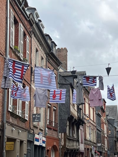 The image captures a street in Honfleur, France, decorated with nautical-themed laundry. Buildings with red brick facades and traditional French architecture line the narrow street. Clothes, primarily striped shirts and banners with red anchors and hearts, are strung across the street, creating a festive atmosphere. Two black umbrella-shaped decorations are also visible, hanging from wires. The sky is overcast, suggesting a cloudy day. The scene evokes a sense of maritime heritage, characteristic of coastal towns like Honfleur.
