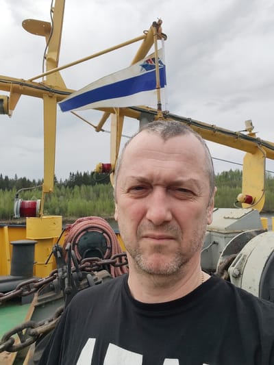 A middle-aged man with short gray hair and a light beard is taking a selfie outdoors. He is wearing a black t-shirt. Behind him, there is a large yellow metal structure with a white and blue flag attached to it. There are also ropes, chains, and other industrial equipment visible, suggesting he is on some kind of boat or industrial platform. In the background, there is a treeline and a cloudy sky. The flag resembles a naval ensign. Given the mention of Alekseevskoe, Russia, it's probable this is a river vessel on the Kama River.