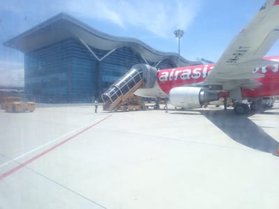 This image depicts an airport tarmac at Cam Ranh City, Vietnam. A red AirAsia airplane is parked, with a mobile staircase attached to its side. The plane's registration number, 9M-RAT, is visible on its wing. Ground crew members are present near the aircraft, attending to operations. In the background, a modern airport terminal with a distinctive wave-like roof design is visible. Yellow airport vehicles, likely baggage tugs or service carts, are positioned on the tarmac. The scene is set under a clear blue sky, suggesting daytime.