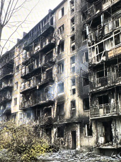 This is a photograph of a multi-story apartment building in Neubrandenburg, Germany, that appears to have sustained significant fire damage. The exterior walls are blackened and scorched, particularly on the upper floors and balconies. Many windows are broken or missing, and the balconies themselves show signs of charring and structural damage. The building is an older style, with multiple levels of protruding balconies on each floor. In the foreground, there is a dense bush or small tree with yellowing leaves, and the ground is littered with debris. Bare tree branches are visible against the sky on the left side of the frame. The overall scene suggests a recent or significant fire event that has left the building in a state of disrepair. There are no visible people or any indications of ongoing activity.
