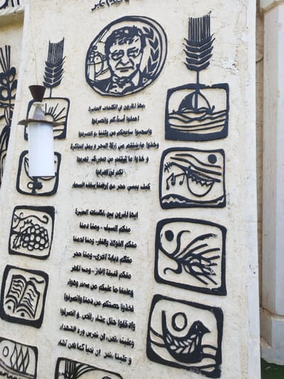 The image shows a wall plaque or monument featuring a portrait of a man surrounded by a wreath. Several stylized symbols appear throughout the monument, including wheat, a boat, musical notes, a bird, and a book. Arabic text is inscribed between the symbols, seemingly a poem or commemorative inscription. The overall style is artistic and memorial. A small light fixture is mounted on the monument.
