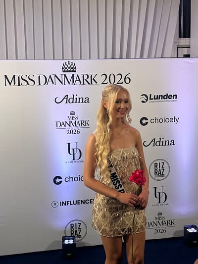 A fair-skinned woman with blonde, wavy hair is standing in front of a banner that reads "MISS DANMARK 2026" and features logos for companies like "Lunden Charlottenlund Travbane," "choicely," "Adina," "RIZ RAZ," and "UNIQ DÉCOR." She is wearing a sequined, strapless dress and a sash that reads "- MISS D." She is holding a red rose. The photo was taken at a location most likely in Skovshoved, Denmark. A lighting fixture is visible in the upper right corner of the frame.