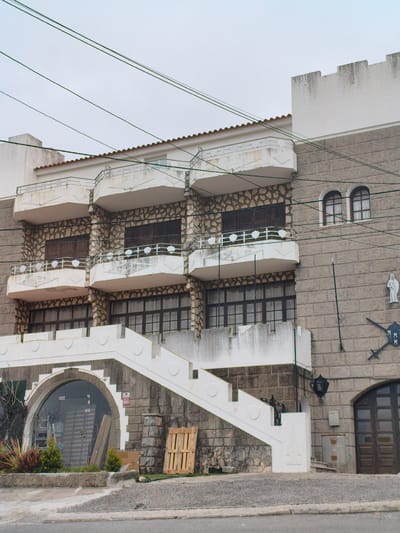 The image depicts a multi-story building with a facade constructed of stone blocks and white plaster. The architecture features arched windows and balconies with decorative railings. A grand white staircase with shield-shaped embellishments leads up to an arched entryway with a glass door. To the right of the staircase, a dark wooden door is visible. In the foreground, there are wooden pallets and some greenery. The sky is overcast. This building is identified as the "Casa dos Peixinhos" in Nazaré, Portugal, a historical landmark that serves as a cultural center.