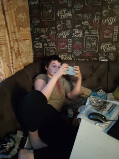 A young person with short, dark hair is sitting on a dark brown sofa, holding a light blue smartphone in both hands and looking at it. They are wearing a grey t-shirt with a red stripe and black pants. The sofa is next to a wall covered in wallpaper with a repeating pattern of cafe-themed text and graphics. To the right of the sofa, a white table is visible, with a newspaper, a computer mouse, and a glass ashtray containing cigarette butts on it. A dark computer keyboard is also partially visible. In the background, a curtain with a floral pattern hangs on the left side of the frame. The overall environment appears to be an indoor living space. The image does not provide specific information to determine the location within Андрэеўшчынскі сельскі Савет, Belarus.