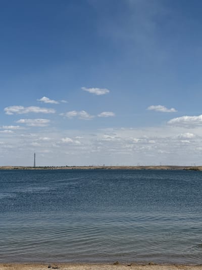 A wide shot captures a serene body of water under a vast blue sky dotted with scattered white clouds. The water, a deep blue, exhibits gentle ripples that become more pronounced as they approach the foreground. The shoreline is composed of light-colored sand and small pebbles, with a few pieces of debris, including a pair of flip-flops, visible near the bottom edge of the frame. In the distance, across the water, a flat, arid landscape stretches out, punctuated by a tall, lattice-like tower and several smaller structures that appear to be power line pylons. The horizon is defined by a hazy line where the land meets the sky. This scene is characteristic of the expansive, open landscapes found near Jezqazğan, Kazakhstan, a region known for its large reservoirs and industrial infrastructure.
