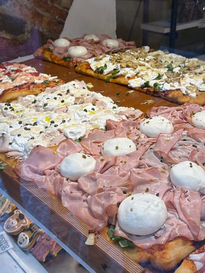 The image shows a close-up view of several rectangular pizzas displayed on a wooden surface, likely in a bakery or pizza shop. The pizzas are topped with various ingredients. In the foreground, a pizza is covered with thinly sliced pink meat, likely mortadella, and several large balls of fresh mozzarella cheese. Scattered on top are chopped green pistachios and capers, with a drizzle of olive oil. Behind this, a pizza is topped with a white creamy sauce, capers, and thinly sliced meats. Further back, another pizza is topped with a creamy white cheese base, green herbs, and what appear to be sliced mushrooms and shavings of hard cheese. The pizzas have golden-brown crusts. In the bottom left corner, a glass display case reveals decorative cookies, one labeled "BISCOTTI." The background shows a brick wall. This scene is consistent with a food establishment in Mozzo, Italy. There are no people visible in the image.