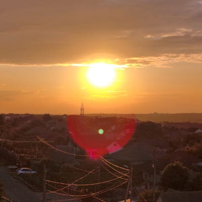 This image captures a sunset over the city of Tohatin, Moldova. The sun, a bright, luminous orb, is positioned high in the sky, casting a warm, orange glow across the scene. Wispy clouds are scattered across the horizon, painted with the colors of dusk. Below the sky, a dense collection of rooftops and trees stretches out, suggesting a residential area. A prominent power line pylon stands tall in the mid-ground, its skeletal structure silhouetted against the vibrant sky. The foreground is dominated by a jumble of power lines, their wires crisscrossing the frame, some illuminated by the setting sun. A silver car is visible on a road in the lower left portion of the image, parked amidst the dense foliage. The overall atmosphere is one of a rural or suburban landscape at the end of the day.