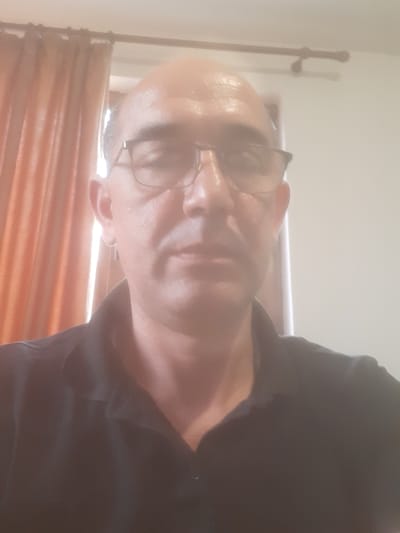 An image depicts a bald, light-skinned individual wearing glasses and a dark-colored garment. The person's head is facing left, tilted slightly downward. Background shows a wooden rod with a clothes rack and an orange window blind. Location: Possibly Dolni Chiflik, Bulgaria.