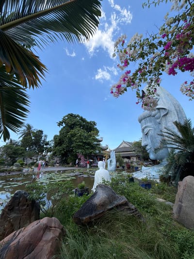 This photograph captures a serene outdoor scene within the city of Hòa Đa, Vietnam. Dominating the foreground is a colossal, light blue statue of a serene face with closed eyes, appearing to be part of a larger deity or figure, adorned with draped fabric. The statue is partially obscured by lush green foliage and large, reddish-brown rocks in the immediate foreground.

Behind the statue, a tranquil pond filled with lily pads and pink and purple water lilies reflects the bright blue sky. Several white Buddha statues are visible, with one meditating figure positioned near the water's edge and a large, open hand statue reaching from the pond. In the middle ground, a dense, mature green tree stands tall, flanked by other tropical vegetation and traditional Vietnamese architecture with gray tiled roofs.

Several people are scattered throughout the scene, observing the statues and gardens. One individual, wearing a long pink dress, stands near the pond, seemingly taking a photograph. Other figures, casually dressed, are also visible in the vicinity. Blooming bougainvillea in shades of pink and white cascades from overhead branches, framing the upper portion of the image and adding a touch of vibrant color. Palm fronds from a nearby tree are visible in the top left corner, contributing to the tropical ambiance. The overall atmosphere is peaceful and spiritual, characteristic of a temple or garden complex.