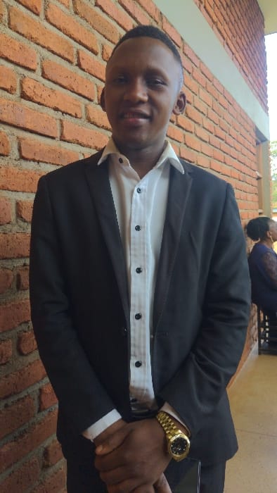 A man wearing a black suit and white shirt stands against a brick wall. He is smiling, with a gold watch on his left wrist. In the background, a blurry figure is visible. The scene is well-lit, capturing the textures of the wall and the man's attire. The photo is taken at an angle.