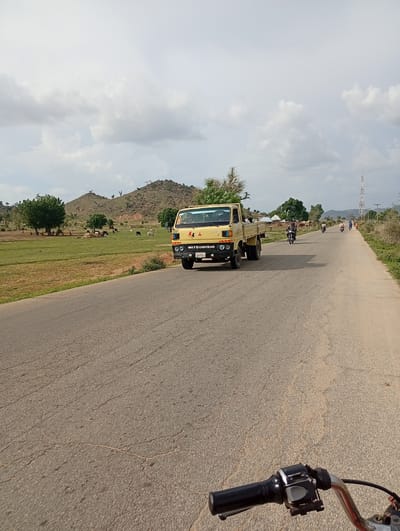 The image shows a road in Shuwa, Nigeria, on a partly cloudy day. A yellow Mitsubishi truck is driving on the right side of the road, with several motorcycles and a few people in the distance. The landscape features rolling hills, green fields, and scattered trees, with a cell tower visible on the horizon. In the foreground, the handlebars of a motorcycle suggest the photo was taken from its perspective. The road is paved and shows signs of wear and tear.