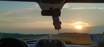 This is a first-person perspective photo taken from inside a vehicle, likely a van or a car, looking out through the windshield. The scene depicts a road trip at sunset in Chýšť, Czechia. The setting is a highway with a few cars visible in the distance. The sky is filled with soft clouds illuminated by the setting sun, creating a warm, golden-orange glow. The time of day is clearly sunset due to the low angle of the sun and the colors in the sky.

The interior of the vehicle is partially visible, including the steering wheel on the left and the dashboard with various controls. A GPS or phone mount is attached to the rearview mirror, suggesting navigation is being used. On the right side of the frame, a person's leg and foot, wearing a light blue sock, are propped up, indicating a relaxed travel posture. The windshield has some dust or smudges, and there are also some faint water streaks, possibly from rain or a recent wash.

The overall mood is peaceful and serene, characteristic of a quiet drive as the day winds down. The landscape outside the vehicle consists of green fields bordered by a fence-like structure on the left and trees on the right, with a bridge or overpass visible in the far distance. The weather appears clear with some scattered clouds. No visible text is present in the image.