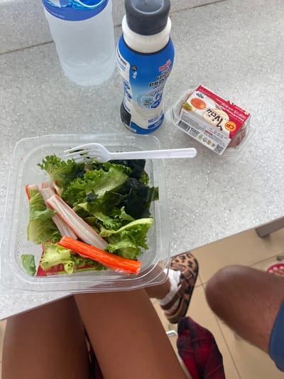 The image shows a close-up, overhead view of food and drinks on a speckled gray countertop, likely in a casual dining or break area. In the foreground, a clear plastic container holds a salad made of green lettuce and what appears to be imitation crab sticks. A white plastic fork rests on top of the salad. To the left of the salad is a clear plastic bottle of water, and further to the left, a blue and white bottle of Meiji High Protein drink. Behind the salad container, there's a small red and white package with Thai text, which appears to be a food product, possibly boiled eggs as indicated by the visible text. The background shows parts of a person's legs, wearing a plaid skirt and leopard-print sandals with white socks, and another person's legs wearing blue shorts. The lighting suggests it's daytime. The overall impression is a casual meal or snack break. The visible text includes "Meiji HIGH PROTEIN," "NO SUGAR LOW ADDED," and details about boiled eggs in Thai.