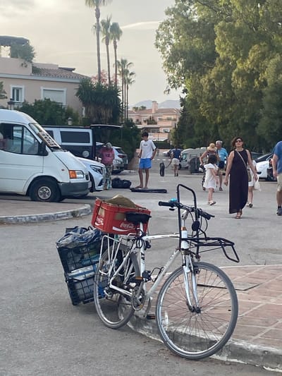 A parked bicycle with a red Coca-Cola crate filled with sand and a black plastic basket containing blankets and possibly other items is positioned on a paved area next to a curb. The bicycle is white and has a front rack. In the background, a white van is parked, and behind it, a dark-colored van with its rear door open. Several people are visible in the mid-ground, including a person in a pink shirt, a young person in a white t-shirt and blue shorts, a woman in a dark dress and sunglasses, and other individuals walking. Palm trees and lush green trees are present, with buildings in the background. The scene appears to be a public area, possibly a parking lot or street in Playa Del Sol-Villacana, Spain, during daylight hours.