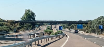 The image shows a highway overpass in Puerto de Santa Cruz, Spain. The sky is clear and blue. The highway has multiple lanes and is bordered by guardrails and vegetation. There are several cars on the road, including a black car and a white car. A large truck is also visible in the distance. There are several road signs, including a blue sign with white arrows indicating two lanes going in the same direction and another blue sign with text indicating destinations and distances. The overpass is a concrete structure with supports visible on either side of the road. The overall scene is typical of a highway in a rural or suburban area.