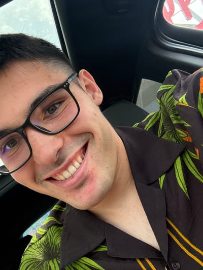 The image captures a close-up, low-angle shot of a young man smiling broadly from within what appears to be a car interior. He has short dark hair with a faded style and is wearing black-rimmed glasses that reflect a subtle purple-blue light. His wide smile reveals his teeth, with some natural yellowing visible on the lower incisors. He has light stubble on his chin and cheeks, and some minor skin redness or blemishes are present.
The man is dressed in a dark-colored, possibly black, collared shirt adorned with a vibrant tropical pattern of green leaves and yellow-orange stripes. Parts of the shirt's collar and an open button are visible around his neck.
The background elements indicate a car interior, with a dark headliner or roof lining visible on the left and top. Through a window in the upper left corner, a bright, overcast sky is discernible, along with a partial red object with white outlines, possibly a letter 'R' or part of a sign. The overall lighting suggests it is daytime. The man's joyful expression is the central focus. The image's context is Radinovo, Bulgaria.