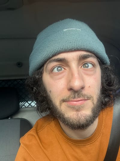The image displays a close-up portrait of a young adult male positioned inside a vehicle. The male has dark, curly hair visible beneath a teal-colored knit beanie, which features a small white horizontal stitch on the front. He wears a dark, medium-length beard and mustache. His light-colored eyes are wide open, with dilated pupils, directed slightly upwards and to the viewer's right. A subtle upward curve is present at the corners of his mouth. He is dressed in an orange crew-neck t-shirt, and a dark seatbelt crosses his right shoulder and chest. Behind him, a dark vehicle headrest and a metal mesh cargo barrier are partially visible, indicating a commercial or utility vehicle interior. The specific location within Marcq-en-Barœul, France, cannot be determined from the visual information provided.