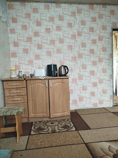 The image depicts an interior scene, likely a kitchen or dining area, in a residential setting in Nimoreni, Moldova. A patterned wallpaper with squares in shades of pink and white covers the wall. In the center of the image, a light-colored wooden cabinet with two doors and a set of three drawers stands against the wall. On top of the cabinet, to the left, are two jars, one with a yellow lid and the other with a darker lid, likely containing food items. Next to them are some small items and a grey mug. To the right on the cabinet are a white toaster and two electric kettles, one black and one silver. A small rug with a bird motif is placed on the floor in front of the cabinet. To the left of the cabinet is a wooden stool with a patterned seat. The floor is covered with a carpet featuring a geometric pattern in shades of brown and beige. A doorway with curtains is visible on the right side of the image. The overall impression is of a functional, lived-in space.