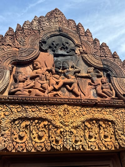 The image shows a close-up view of intricate sandstone carvings on the exterior of Banteay Srei temple in Cambodia. The carvings depict figures in a narrative scene, with multiple individuals engaged in dynamic poses. Some figures appear to be in combat or engaged in a ritual, with one figure on the left seated with hands clasped and another figure on the right appearing to be in motion, reaching out. The central figures are intertwined, suggesting a moment of interaction or conflict. Above this main scene, smaller figures are visible within a darker carved panel, and the entire composition is framed by elaborate decorative elements, including floral motifs and scrollwork. The sandstone is reddish-orange in color, showing signs of age and weathering. The sky above is partly cloudy. The lower portion of the image reveals a highly decorative frieze with more detailed patterns and smaller sculpted elements.