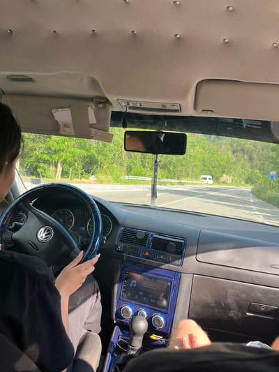 The image shows the interior of a car, looking forward. A woman is driving, with her hands on the steering wheel. The dashboard has a blue, patterned trim. Outside the car, there is a road with trees and hills in the background. A white van is visible in the distance. The scene appears to be captured on a sunny day, likely in the city of Leshtak, Bulgaria.