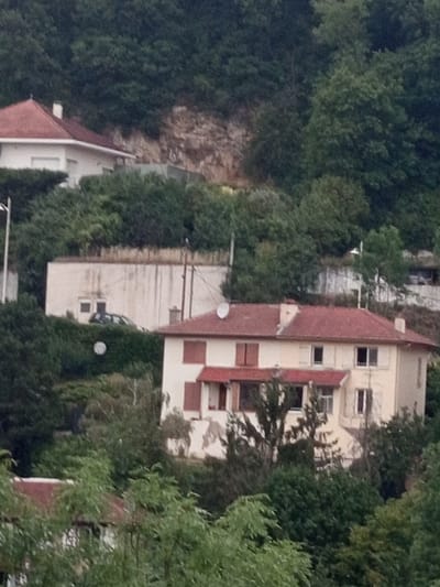 This is an image of a hillside residential area in Nancy, France, featuring multi-story buildings nestled amongst dense green foliage. The buildings appear to be a mix of older and newer construction, with some exhibiting a more traditional architectural style with red tiled roofs and light-colored facades. One building has a prominent red roof and white walls, with visible windows. Another, taller building on the right side of the frame, shows a more modern design with distinct bands of red and white on its exterior. The greenery surrounding the structures is lush and appears to be mature trees and bushes, suggesting a natural or semi-rural setting within the urban landscape. There are no visible people or specific activities taking place. The lighting suggests it is daytime, likely with overcast or diffused sunlight, as there are no strong shadows. The overall impression is of a quiet, residential area integrated with its natural environment.