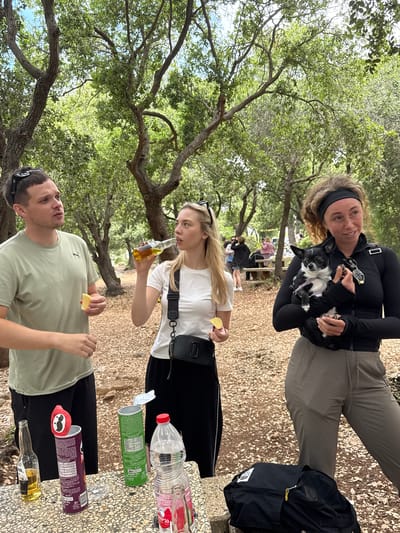 Nesher, Israel. A group of three individuals are gathered outdoors at a picnic table. One individual is holding a small dog. Two individuals are holding slices of lemon. One individual is drinking from a bottle. An opened container of Pringles potato chips is on the table.