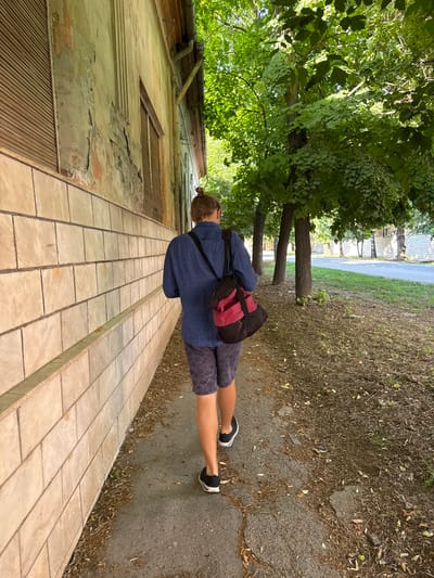 A single individual is observed from behind, walking on a paved sidewalk in Becej Municipality, Serbia. The person has light-colored hair tied in a bun and is wearing a dark blue collared shirt, dark patterned shorts, and black athletic shoes with white soles. A red and black shoulder bag is slung over their right shoulder. To the left, an old building features a lower section clad with light-colored rectangular tiles and an upper wall with significant peeling paint and exposed plaster, revealing underlying material. Several windows on the building are covered with light-colored wooden slat shutters, and a gutter system is visible along the roofline. To the right of the sidewalk, a narrow strip of dry ground is covered with fallen leaves and debris. Beyond this, a line of mature trees with dark trunks and dense green foliage runs parallel to the sidewalk. Further in the background, a paved road is visible, with more trees and distant structures on the opposite side. The scene is illuminated by bright daylight, casting some shadows.