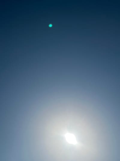 A vertical, upward-facing outdoor shot of a clear blue sky. The sun is visible in the lower right portion of the image as a bright white orb with rays of light extending outwards, creating a lens flare effect. In the upper center of the image, a small, bright teal-colored dot is visible against the blue sky. The image was taken in Vangaži, Latvia.