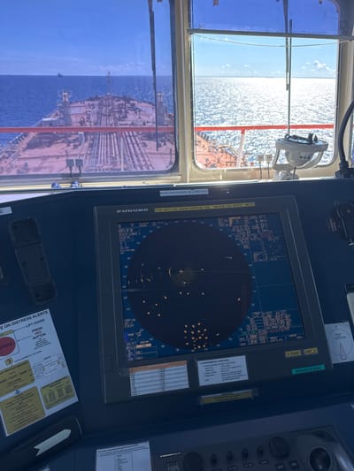 The image shows the bridge of a large ship, likely a tanker, with a view of the sea and the ship's deck through the windows. The primary focus is on the ship's navigation equipment, specifically a Furuno radar display. The radar shows multiple contacts, indicating other vessels or navigational aids. The display also shows various operational parameters and environmental information.

To the left of the radar, there is a panel labeled "ON DISTRESS ALERTS" with instructions for activating Mayday and other distress signals. Below this, there is a form for recording distress and communication information, including fields for Mayday, Callsign, Name, and Position.

In front of the radar, there are various controls and displays, including a joystick-like device. A compass is visible to the right, mounted near the window.

The environment outside the bridge is a sunny day with a calm, blue sea. The deck of the ship is visible, with various pipes and structures characteristic of a tanker. Other ships can be seen in the distance on the horizon.

There is no indication of the city of Staroe Garkolovo, Russia, in the image or any of the provided crops. The scene is clearly on a ship at sea.
